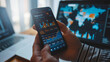 © MP Studio - close-up of a hand holding a smartphone with stock market data on the screen, with a laptop in the background displaying additional financial charts
