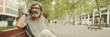 © Andrii Nekrasov - Laughing middle-aged man with gray hair and beard wearing casual clothes sits on bench and uses mobile phone, Panorama. Mature gentleman in eyeglasses talking on smartphone
