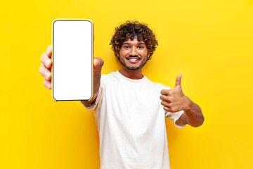 cheerful indian man showing blank smartphone screen and like on yellow isolated background, curly guy advertising phone online and recommending