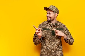 Wall Mural - Ukrainian army soldier in camouflage uniform pixie points with his hands to an empty space on a yellow isolated background, Ukrainian military cadet shows and advertises copy space