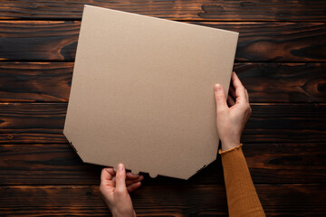 Pizza closed carton box in caucasian hands on natural dark wooden table flat lay mockup with copy space