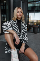 Wall Mural - Fashion beautiful young woman model in fashionable clothes with a zebra print dress and white sneakers sits in the city near a building