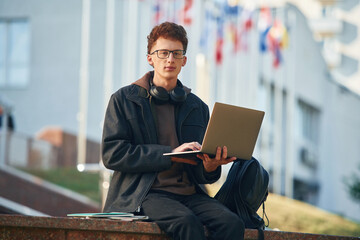 Wall Mural - Guy is sitting and using laptop. Young student is outside the university, in black clothes