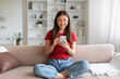 © Prostock-studio - Delighted young asian woman browsing on smartphone while comfortably sitting on couch