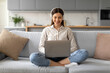 © Prostock-studio - Smiling young woman with headphones using laptop on sofa