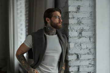 Wall Mural - Successful handsome hipster man with tattoo and beard in fashion clothes with a white T-shirt and sweater stands near the window in the room