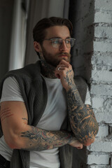 Wall Mural - Male indoors portrait of a handsome hipster businessman guy with a beard and hairstyle with tattoos with glasses in a white T-shirt with a sweater stands near a brick wall by the window