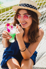 Wall Mural - attractive young woman in blue dress and straw hat wearing pink sunglasses drinking cocktails