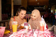 © Connect Images - Two young female friends one wearing a Hijab sit together in a restaurant playing with their phones and showing each other photos