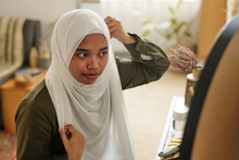Young Arabic Woman Sitting By Toiletry Table And Looking In Mirror While Getting Dressed And Covering Her Head With White Headscarf