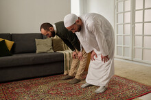 Muslim Men Keeping Eyes Closed And Hands On Knees While Standing On Rug At Home, Bending Forwards And Performing Namaz Prayer