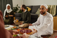 Young Muslim Man Taking Plate With Sweet Homemade Pie From Hands Of Woman While Sitting On The Floor Against His Brother And Wife