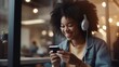 Positive african american teen listening to music on headphones while browsing mobile phone checking playlist sitting in studio with blurred background