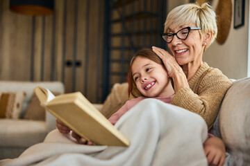 Wall Mural - Grandma and grandchild spending time together, covered in a blanket, reading a book together.