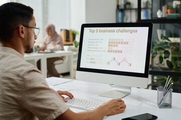 Leinwandbilder - Serious young businessman organizing work while sitting by desk in front of computer monitor with top five business challenges on screen