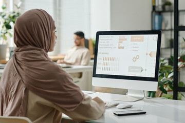 Leinwandbilder - Young Muslim businesswoman in hijab sitting by desk in front of computer screen with statistic data and analyzing it against male coworker