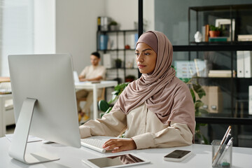 Leinwandbilder - Young female office worker in hijab sitting by workplace in front of computer screen and analyzing data in coworking space