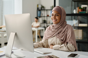 Leinwandbilder - Happy young female analyst or broker in hijab looking at computer screen with smile while sitting by workplace and analyzing data