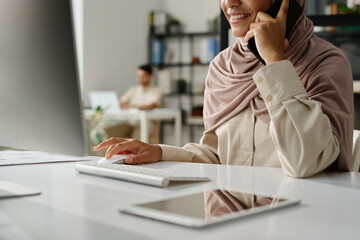 Leinwandbilder - Cropped shot of young smiling office manager in hijab talking on mobile phone and clicking mouse while sitting in front of computer