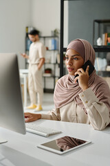 Leinwandbilder - Young confident manager in hijab speaking on smartphone in front of desktop computer screen while sitting by workplace in office