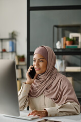 Leinwandbilder - Muslim female manager in hijab holding smartphone by ear while sitting in front of desktop computer in office and talking to someone