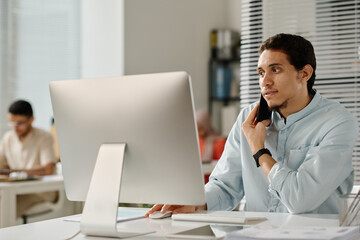 Leinwandbilder - Young confident male manager holding smartphone by ear while sitting in front of desktop computer in office and phoning colleague