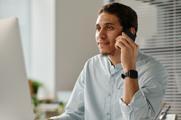 Leinwandbilder - Young male employee talking to someone by mobile phone while sitting in front of desktop computer and scrolling through online data