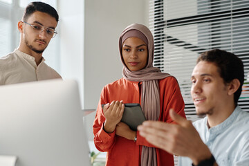 Leinwandbilder - Young businesswoman in hijab with tablet in hands looking at computer screen and listening to explanation of data of male colleague