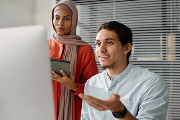 Leinwandbilder - Young confident Muslim male analyst explaining data on computer screen to female colleague in hijab during presentation of project points
