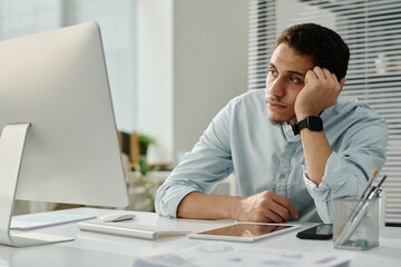 Leinwandbilder - Young bored businessman sitting by workplace in front of desktop computer and looking at screen while waiting for page uploading