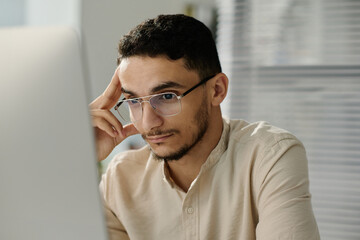Leinwandbilder - Young Muslim male white collar worker in eyeglasses looking at computer screen while analyzing online data or watching video