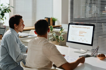 Leinwandbilder - Two young male managers looking through online statistic data on screen of desktop computer while sitting by workplace in office