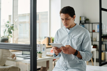 Leinwandbilder - Young serious male analyst or broker with tablet looking through online data while standing in front of camera in coworking space