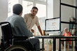 © pressmaster - Young serious male office manager looking at his colleague in wheelchair during discussion of project points or preparation of presentation