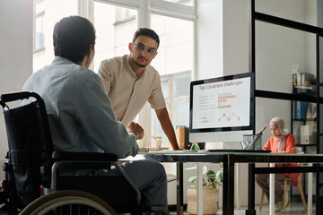 Young serious male office manager looking at his colleague in wheelchair during discussion of project points or preparation of presentation