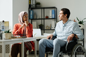 Leinwandbilder - Young Muslim male office manager with disability sitting in wheelchair and looking at female colleague in hijab during discussion