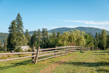 Canvas Print - Rural landscape with mountains on the background, calm summer day. Small path near the wooden fence, green grass, trees and hill, bright blue sky, the sun shining