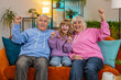 © Andrii Iemelianenko - Happy Caucasian grandparents and granddaughter watching TV shouting, celebrating success, winning, goal achievement good news, victory sitting on sofa in living room. Family clenching fists at home.