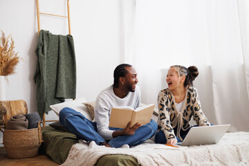 Laughing young multiethnic couple looking at each other while using laptop and reading book on bed at home