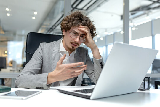 Young male professional with curly hair shows frustration while working on laptop in a modern office environment.
