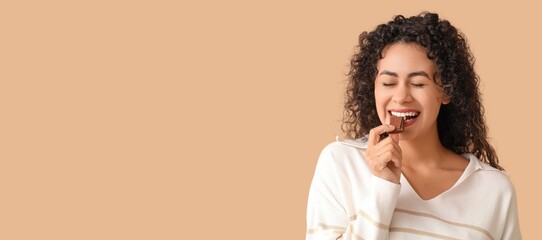 Wall Mural - Beautiful young African-American woman eating sweet chocolate on beige background with space for text