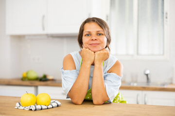 Wall Mural - Young positive housewife in apron posing in modern kitchen