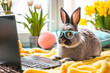 A rabbit wearing glasses sits on a blanket next to a laptop. This image can be used to represent technology, productivity, or studying