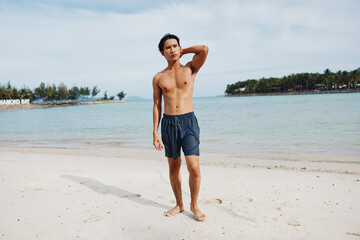 Wall Mural - Smiling Asian Man Enjoying Beach Vacation: Muscular Torso and Happy Smile against Tropical Island Background