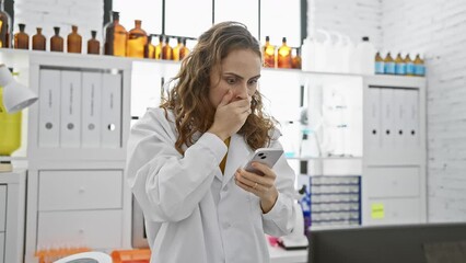 Wall Mural - Beautiful uniform-wearing woman in lab, shocked and afraid, mouth covertly covered by hand in surprise over smartphone misstep