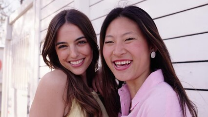 Wall Mural - Close up cheerful pretty Caucasian and Asian women posing looking smiling at camera pulling hair away from face. Two happy friends enjoying laughing together outdoor. Concept of friendship in gen z