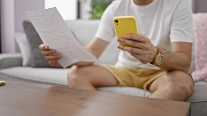 A mature man with grey hair uses a smartphone while sitting on a sofa indoors, holding paperwork and wearing a white shirt and yellow shorts.