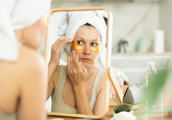 relaxed woman in her middle age using under eye patches for skincare sitting at the mirror in home e