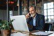 © FrameFlow - Happy middle aged business man talking on mobile phone working on laptop in office. Professional businessman executive wearing suit making call on cell using computer communicating, Generative AI