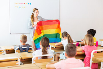 Wall Mural - Schoolchildren sitting in classroom and listening to female teacher. She holding rainbow flag in hands and talking about minorities.